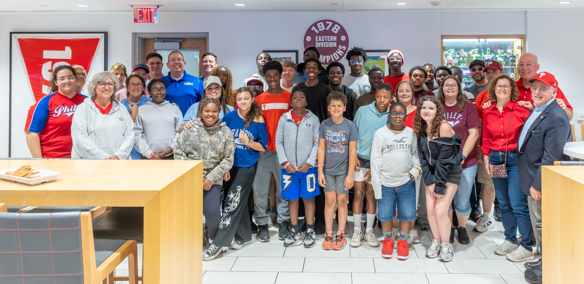 Smiling group of children and adults standing together in a room at a Phillies game.