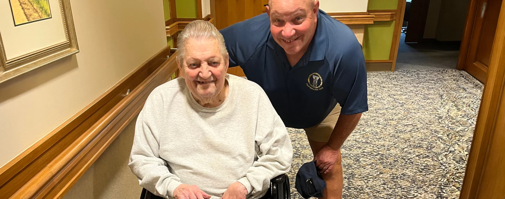 Larry and Don Smiling men, one of whom is seated in a wheelchair, together in a hallway.